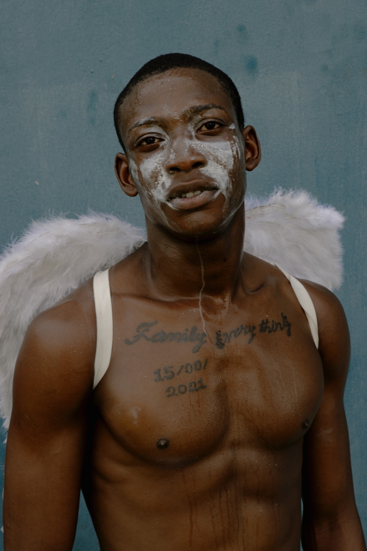 A man wearing white feathery wings with silver paint on his face looks at the camera.