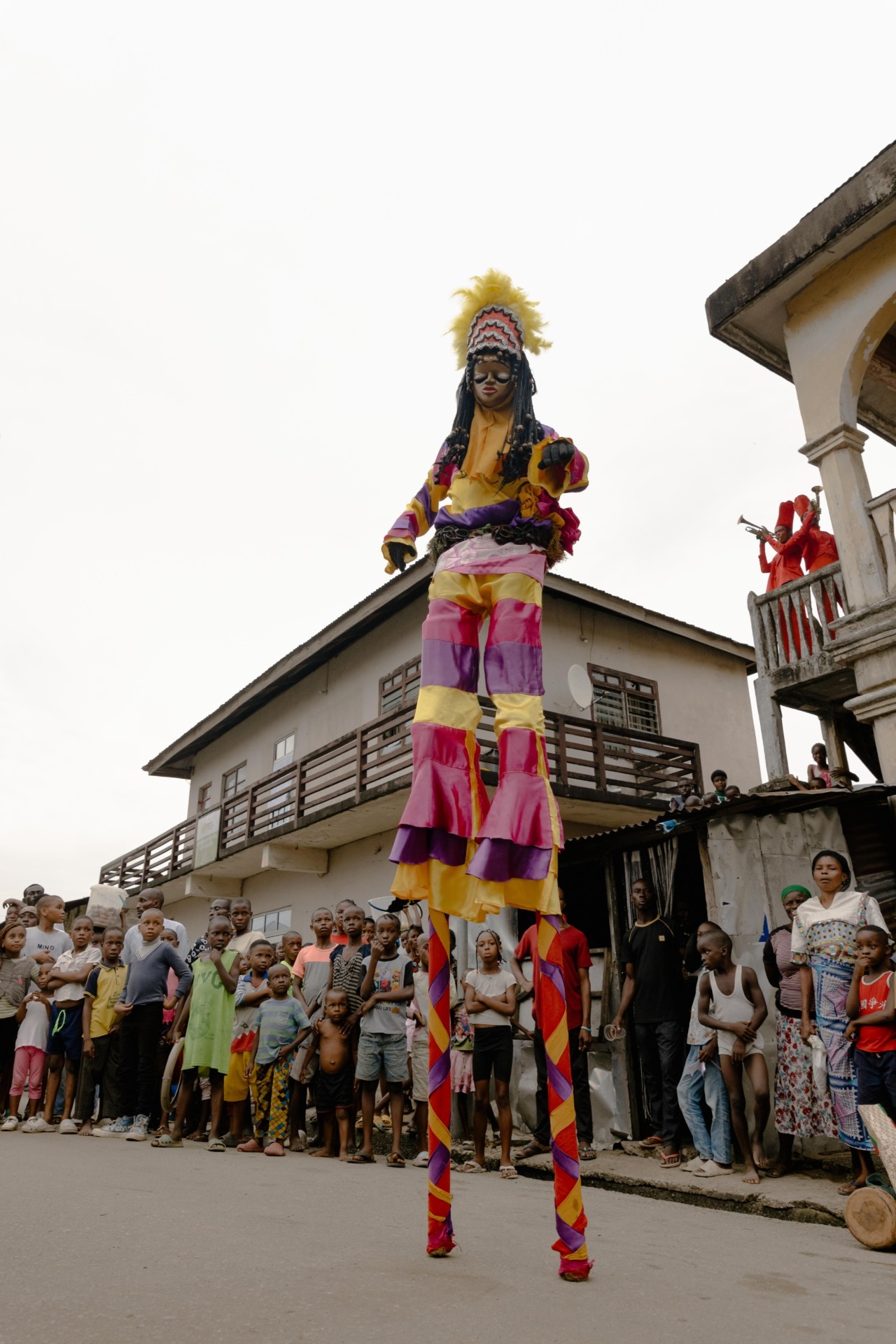 A person dressed in a colorful costume stands on stilts while a crowd of children look on in the background.