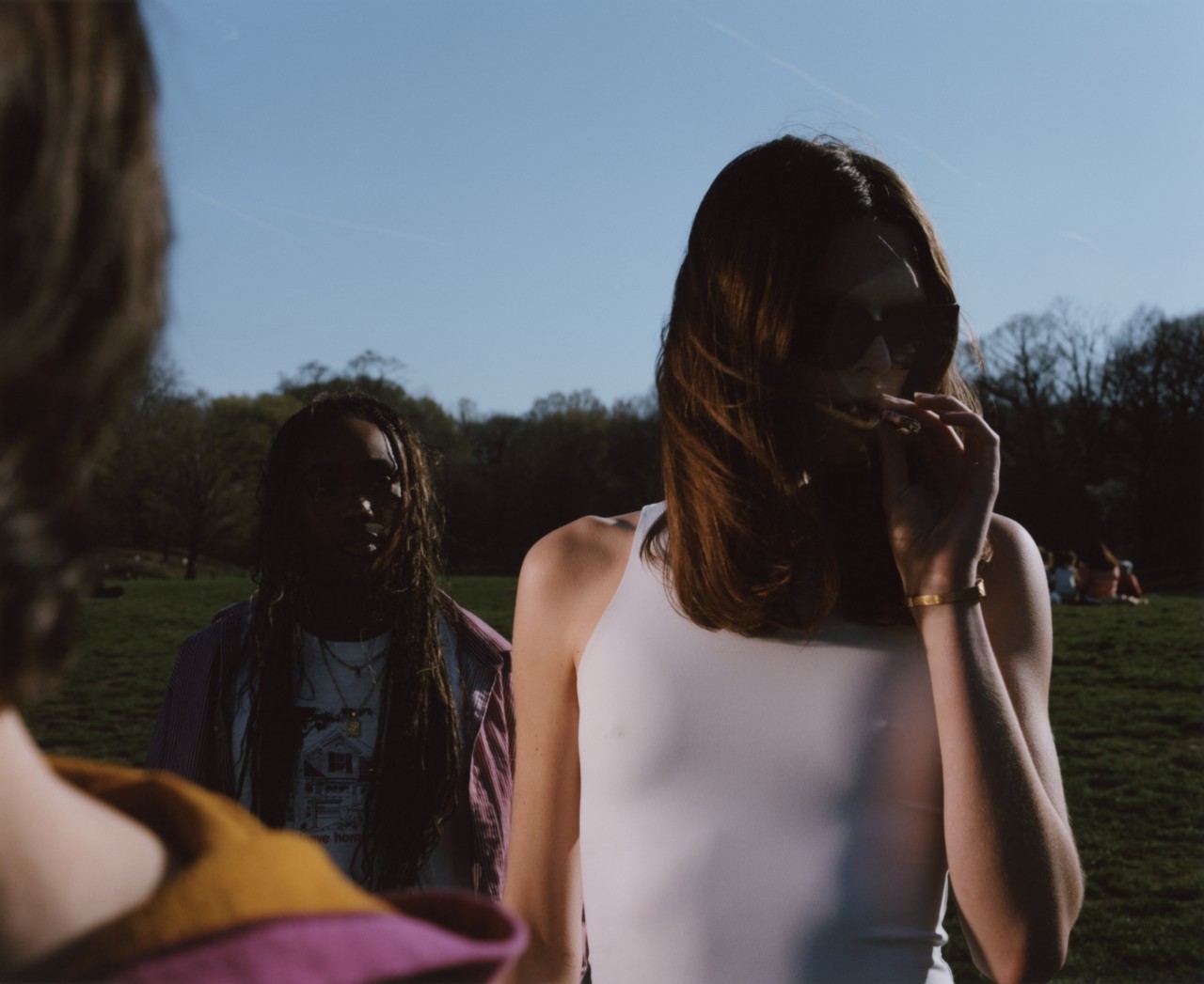 Two teenagers stand in a park, one in the forefront smoking and one in the background watching