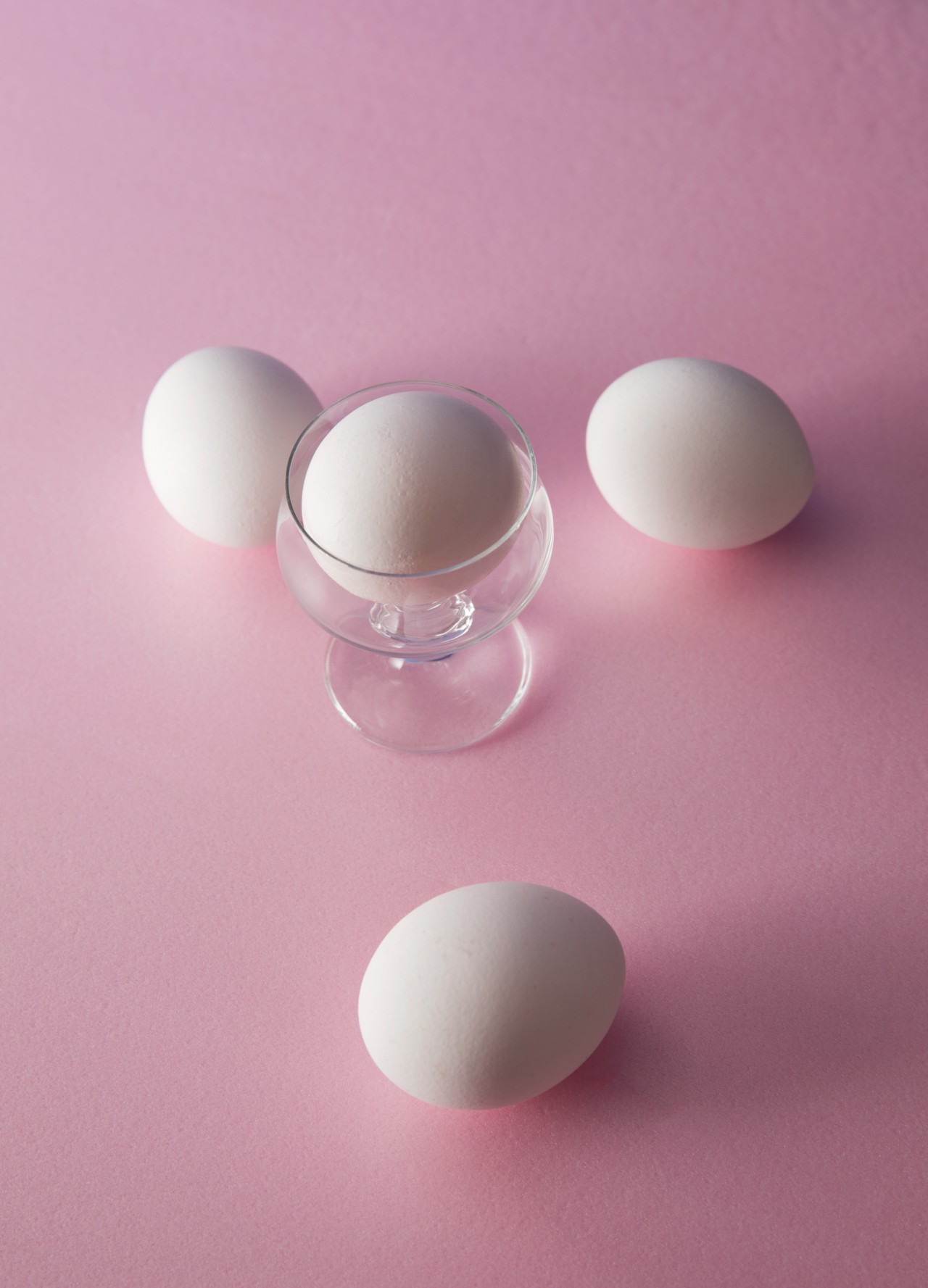 Three eggs sit on a bright pink background. A fourth egg sits inside a small glass in the center.
