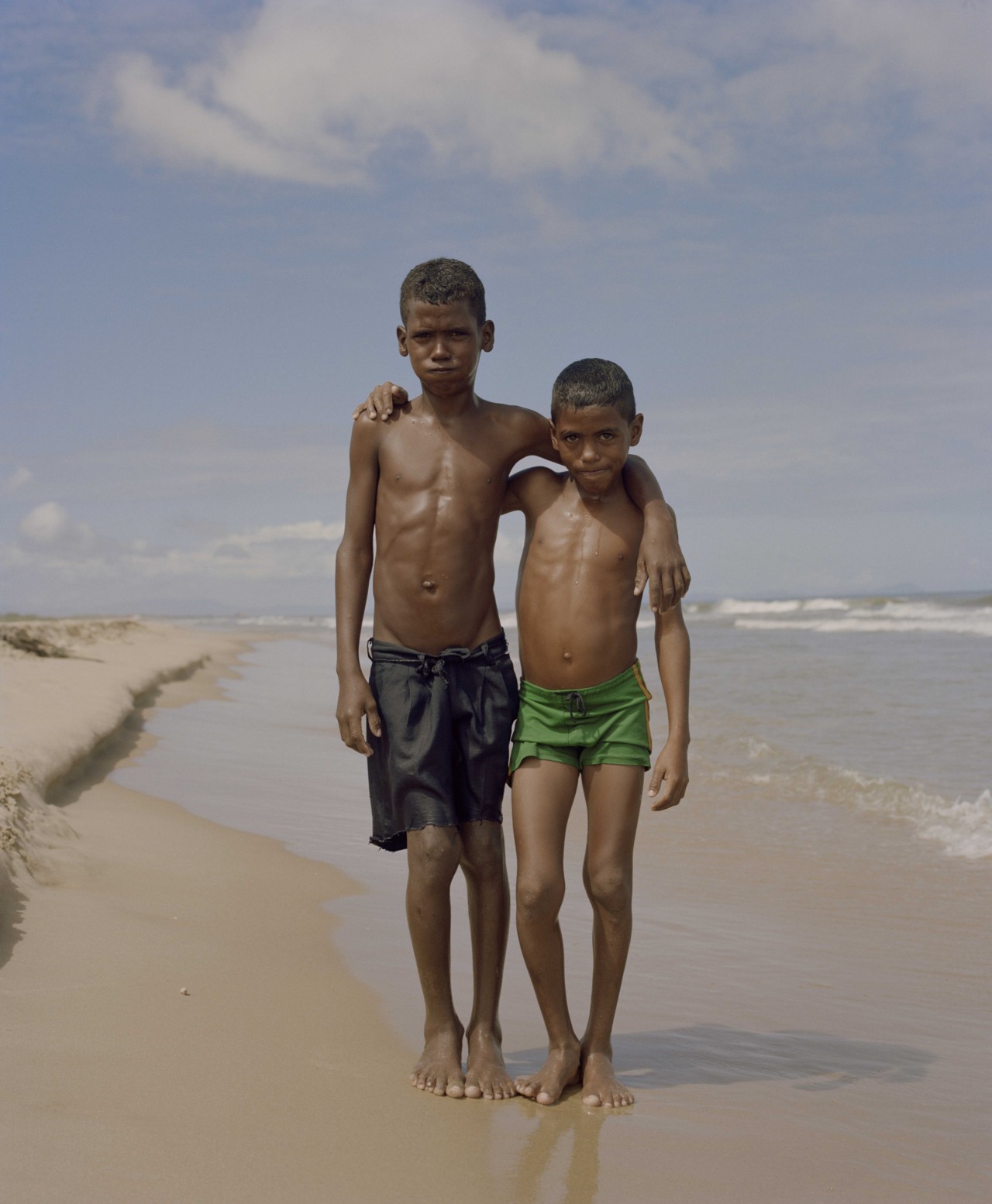 Two Venezuelan boys stand on a beach with their arms around each other.