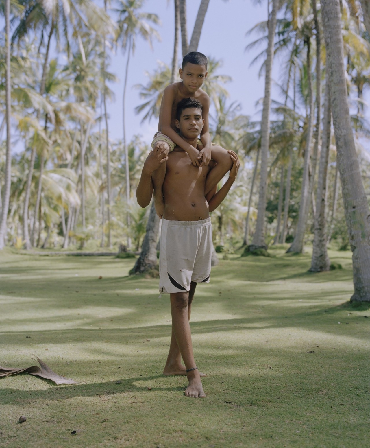 A Venezuelan adolescent carries a young boy on his shoulders, standing on a bed of grass amidst tropical trees.