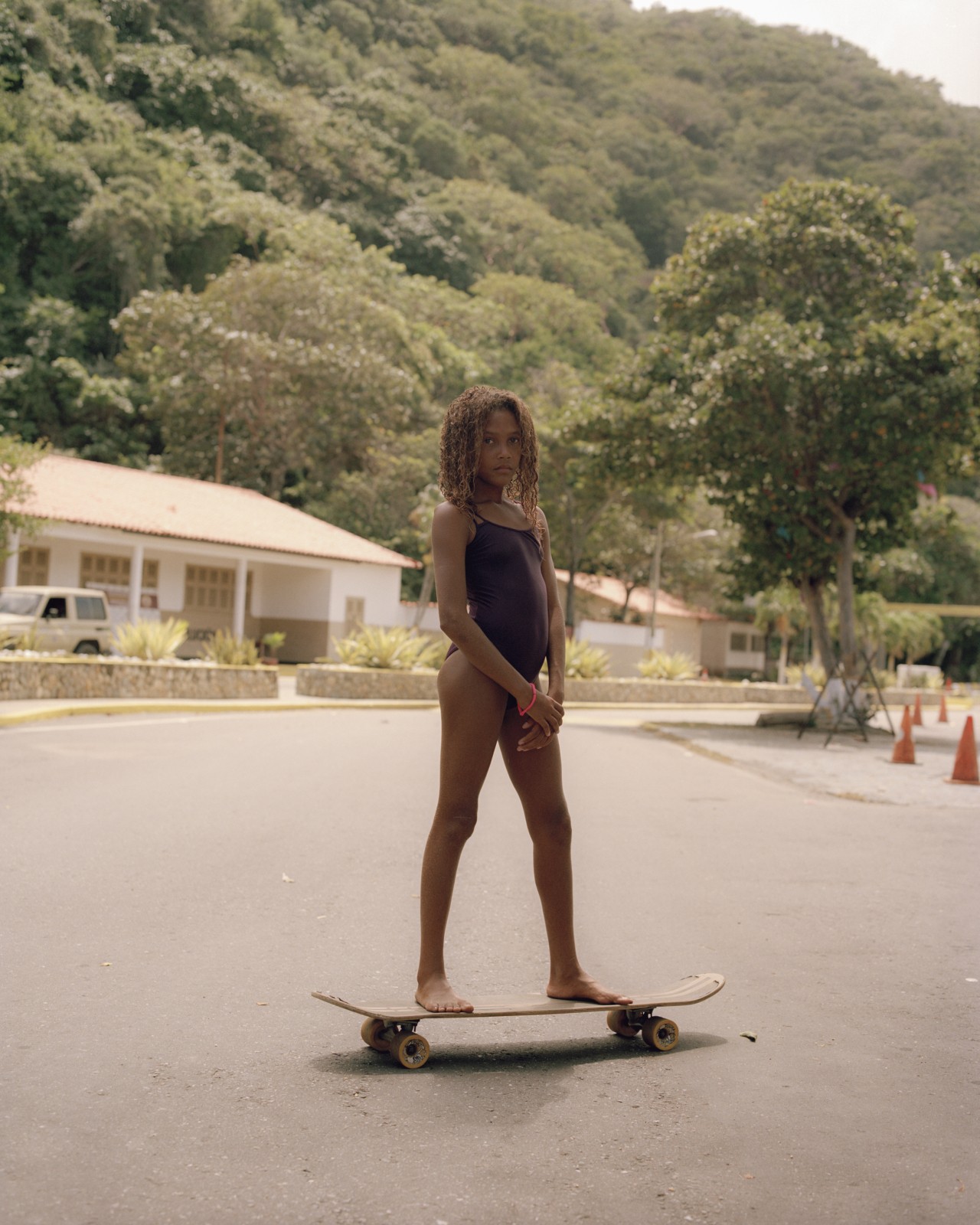 A Venezuelan girl stands on a skateboard in the middle of the street. There is lush greenery in the far distance behind her.