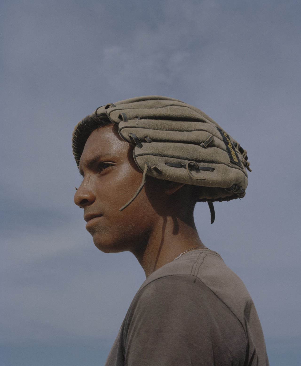A young man from Venezuela man stares in distance with a baseball mitt oh top of his head.