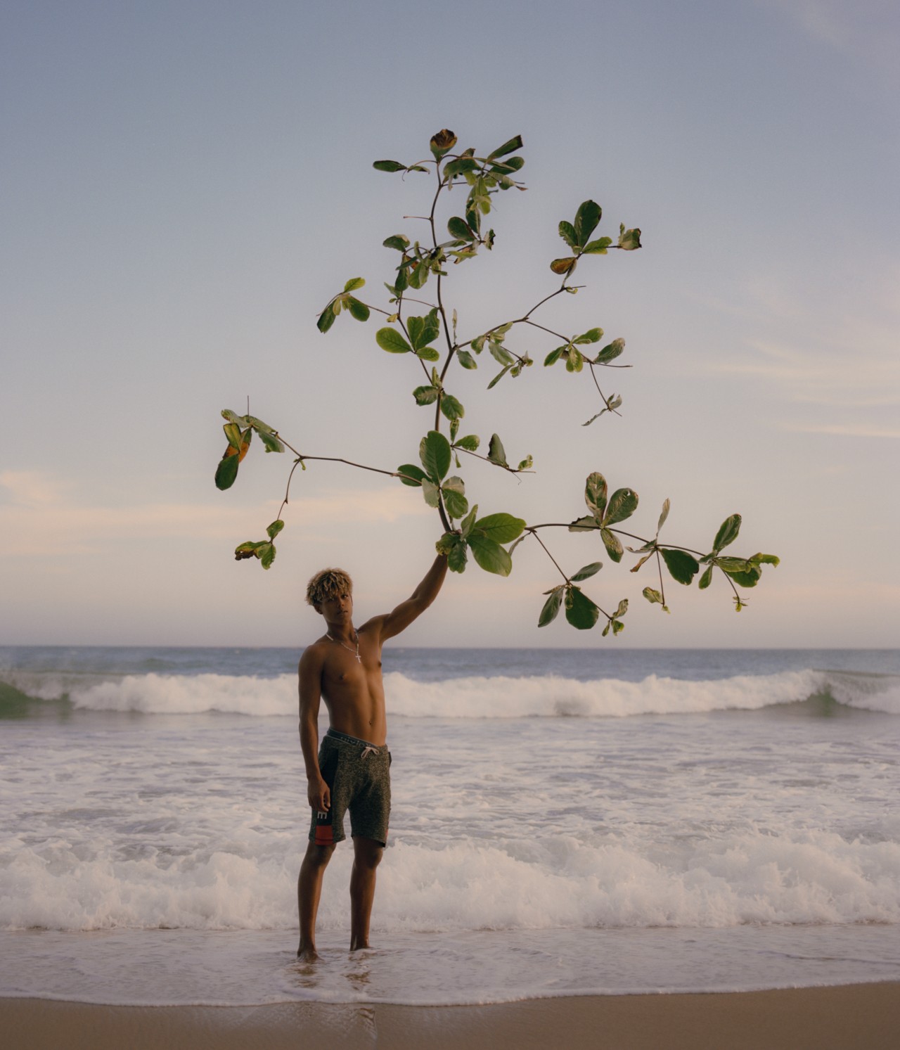A young man from Venezuela stands on the shore of a beach holding a tree branch.