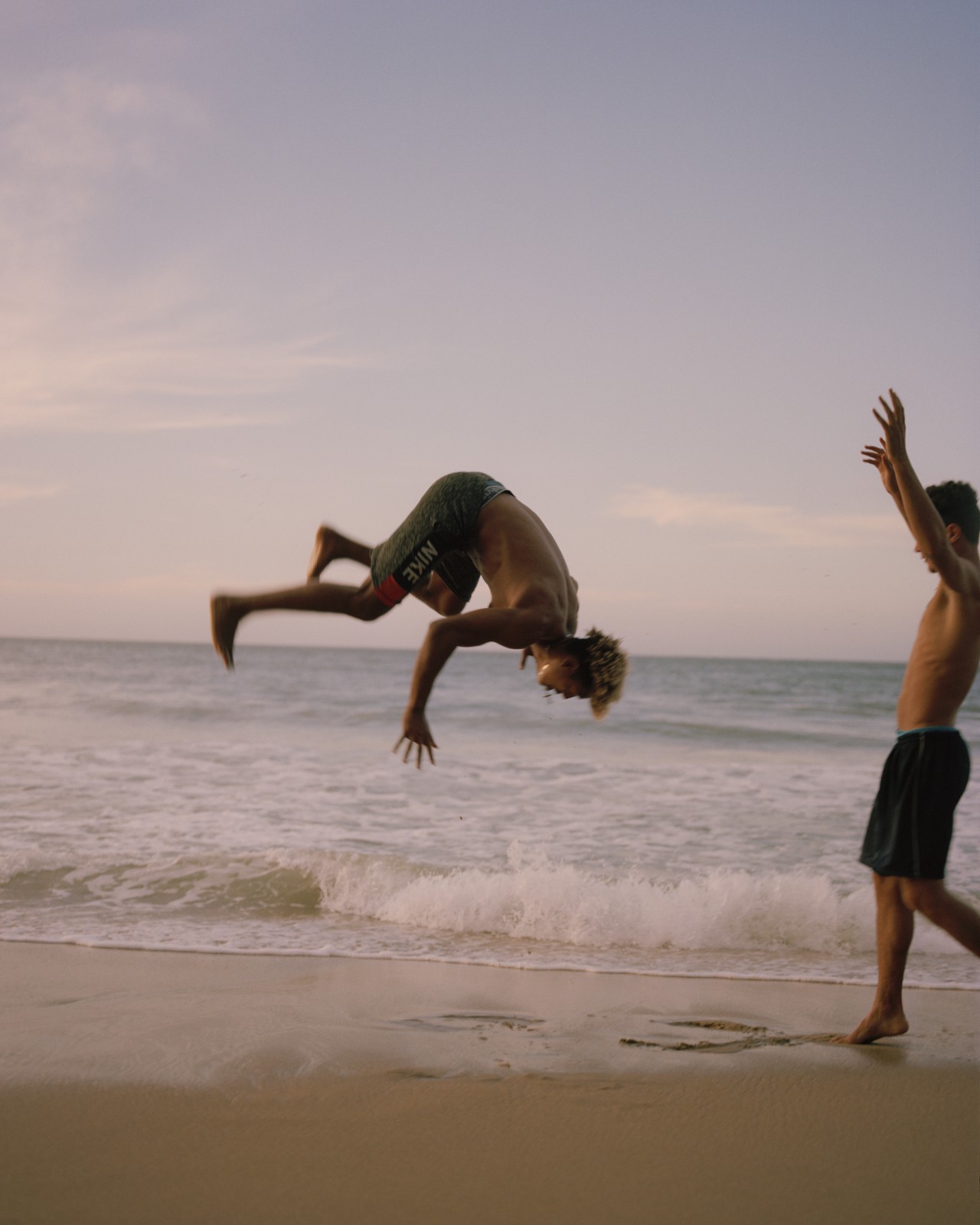 Two Venezuelan youth are playing on the beach. One is in the air mid-flip while the other one cheers on the side.
