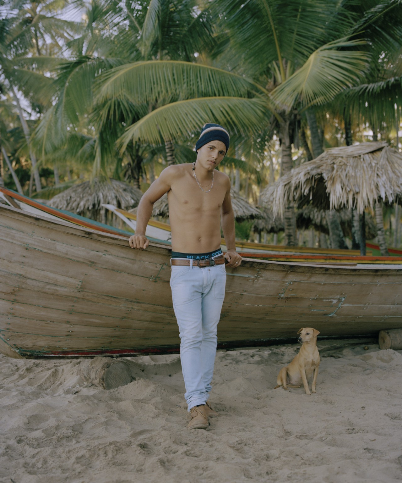 A young man from Venezuela leans against a large boat on the beach. A small dog is sitting next to him and there are tropical trees in the background.