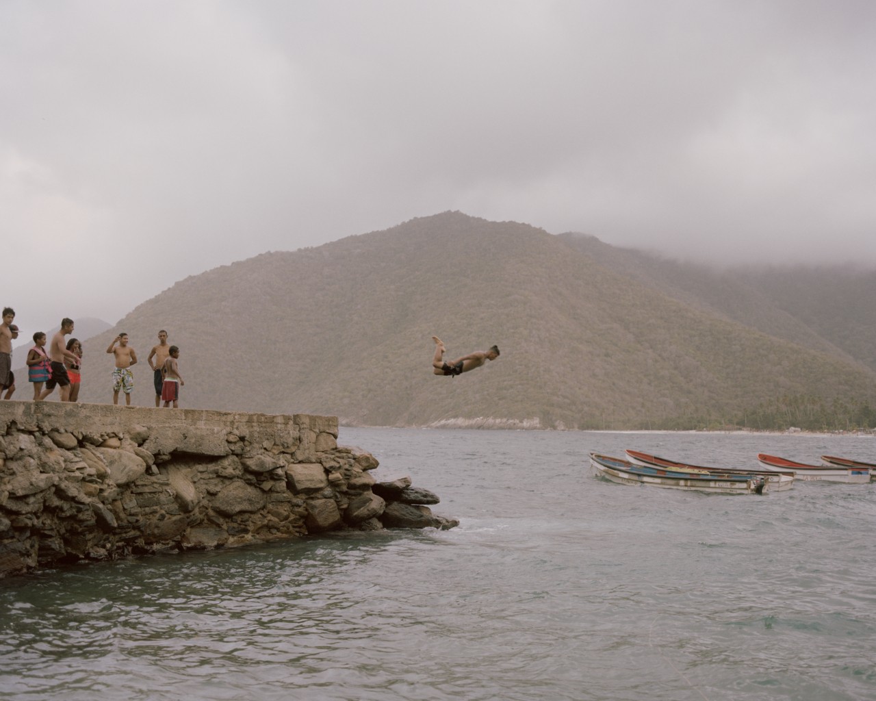 Several Venezuelan youth gather on top of a small cliff. One person is in mid-air, diving into the water.