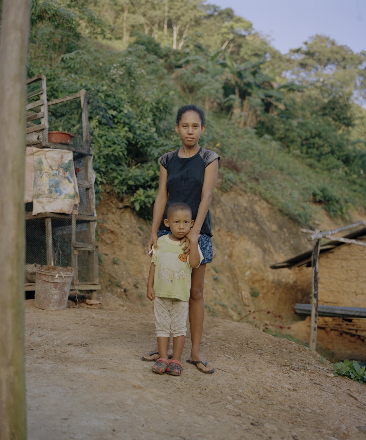 A Venezuelan girl stands on small dirt hole while holding a small Venezuelan boy in front of her. A lush green hill is in the background.