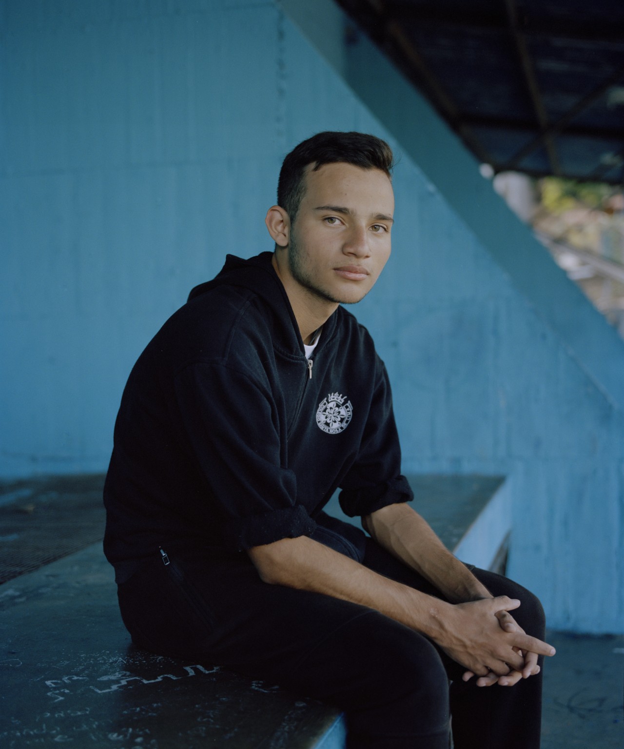 A young man from Venezuela is sitting down on a bench, looking into the camera.