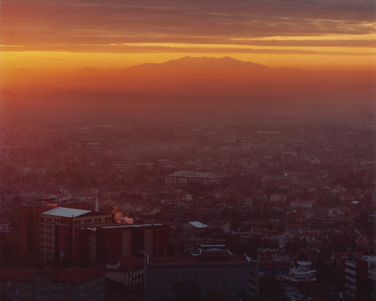 A smoky city landscape meets a hazy orange horizon with a mountain in the distance.