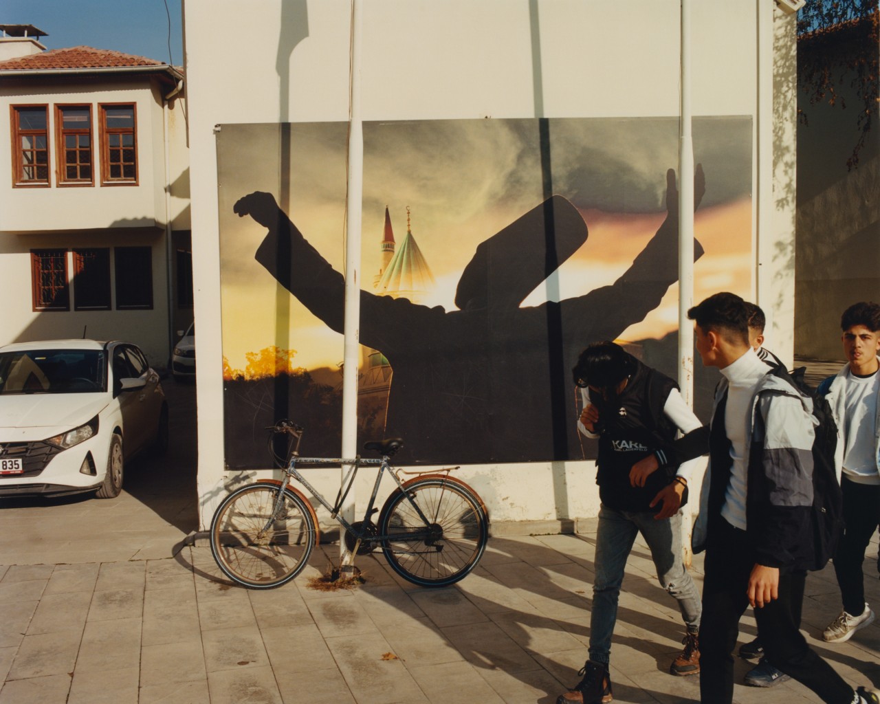A group of young men walk by a large poster of a whirling dervish pasted on a white wall. A bicycle is leaning against the wall and house with a car in the front is on the left side of the wall.