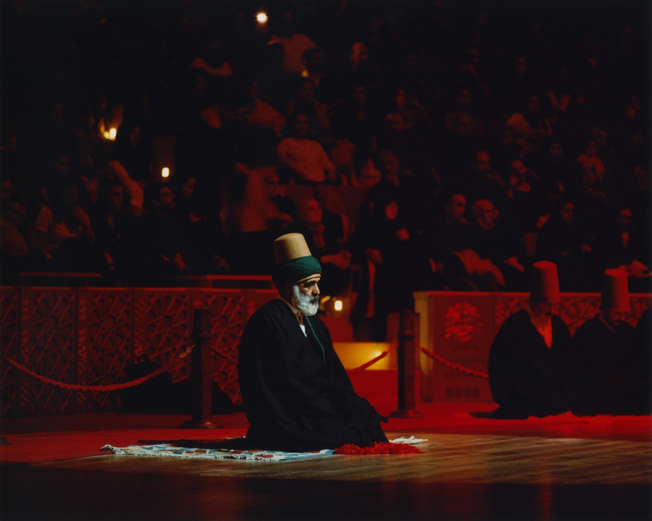An older man sits on the floor facing away from a crowd at a sema ritual.