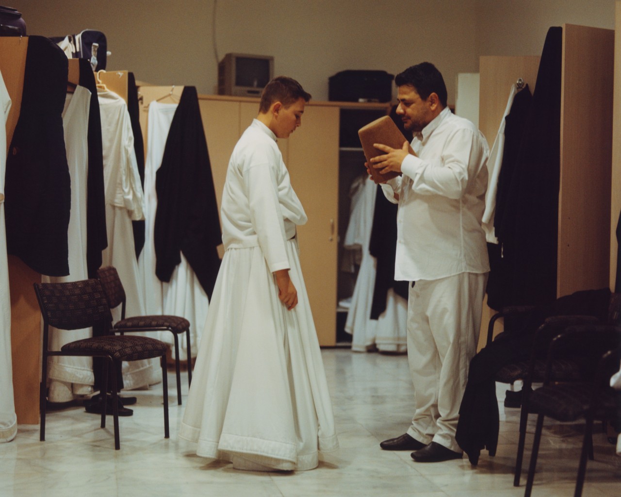 A young male whirling dervish waits as an older man is about to place a brown hat on his head.