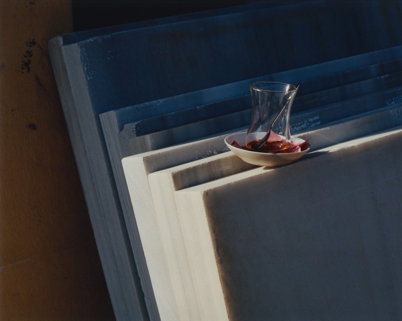 A glass cup sits on a small saucer on top of several large slabs of concrete that are leaning on a wall.