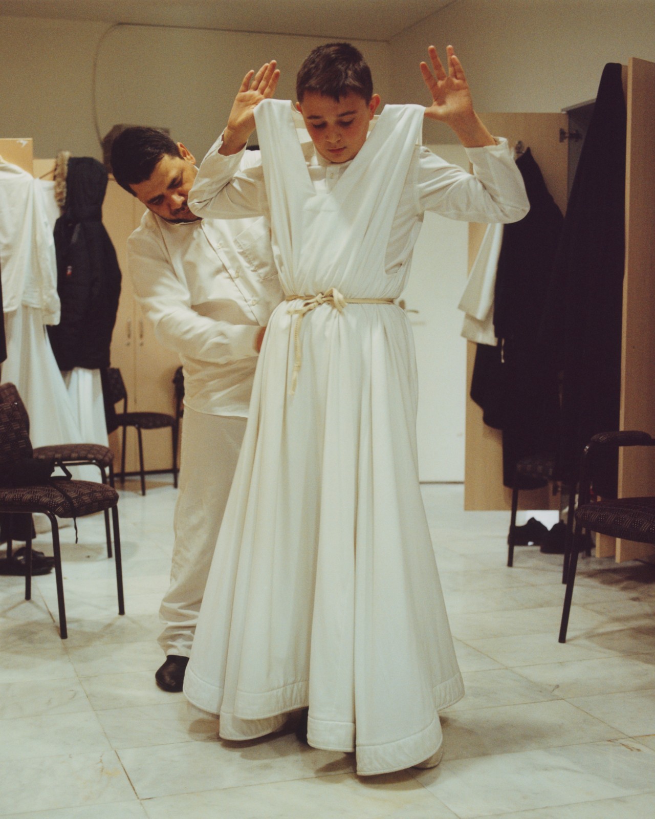 An older man is helping a young male whirling dervish with his clothing.