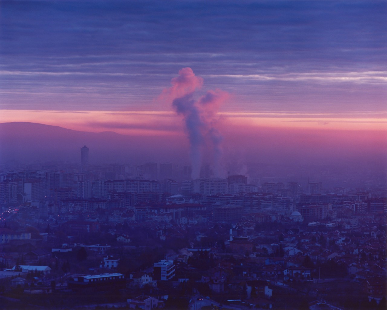 A city landscape shows pinkish blue skies in the background and bellowing smoke in the distance.
