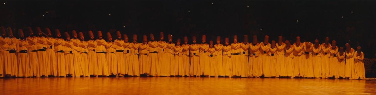 Whirling dervishes stand in line facing the audience, their arms folded across their bodies.