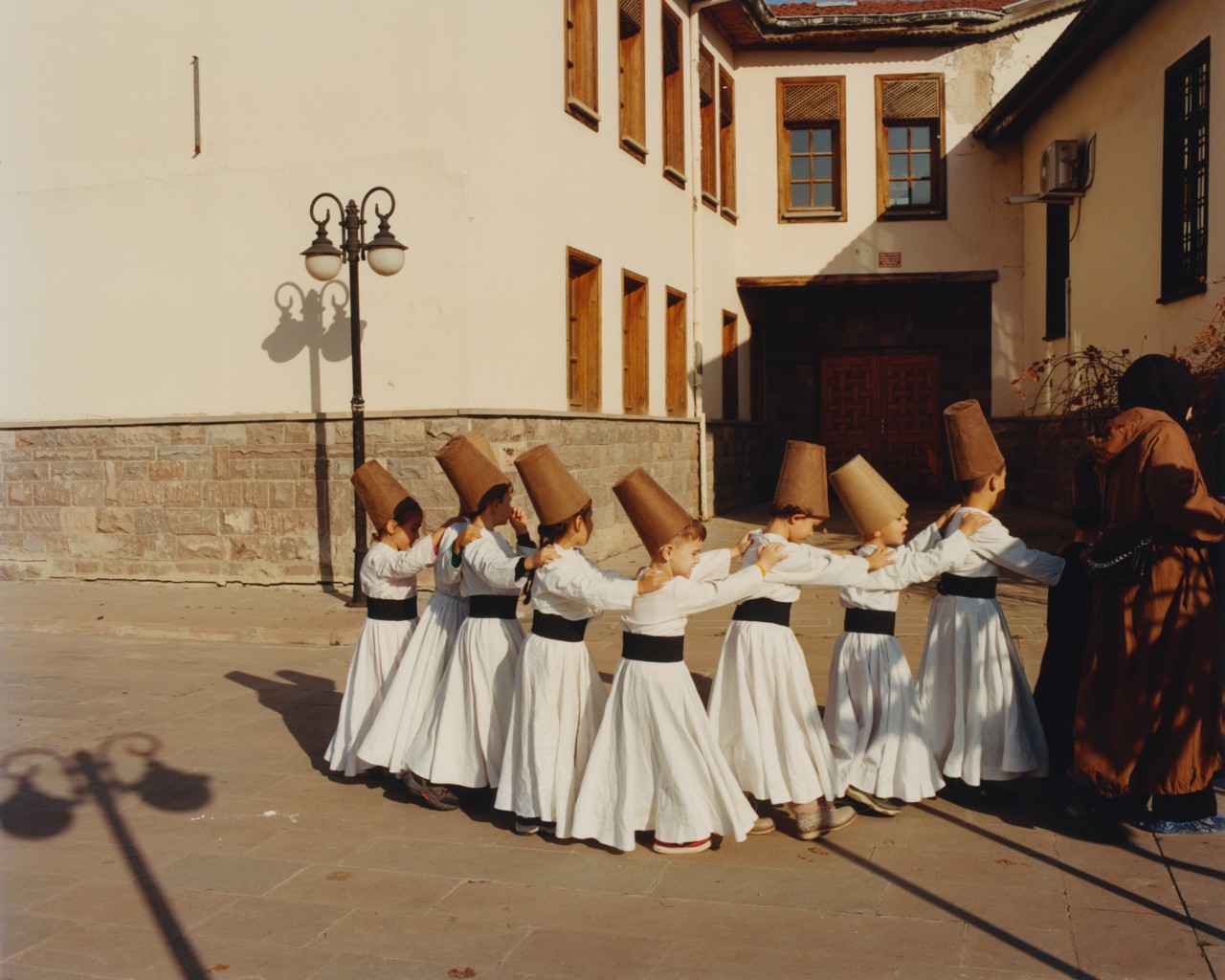 Whirling dervish children draped in traditional garb walk in a single file line, holding onto each other's shoulders.