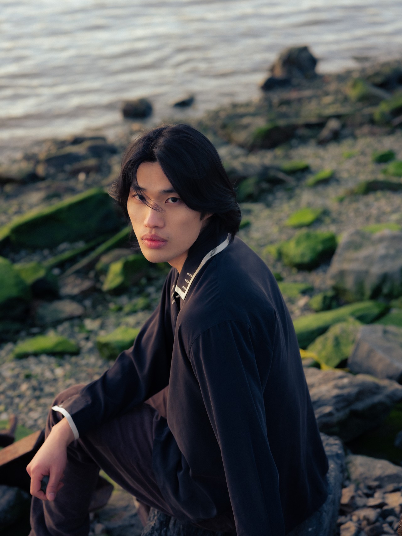 An Asian-American male sits on a rocky shoreline