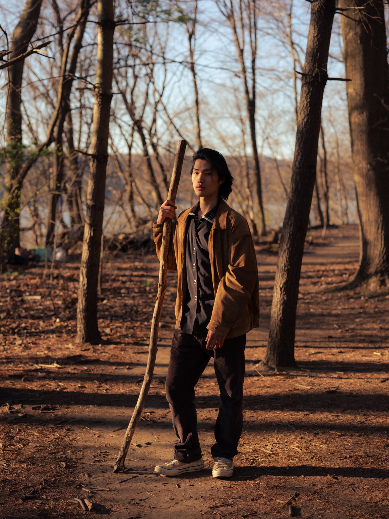 A young Asian-American man stands in the middle of a forest holding a large stick.