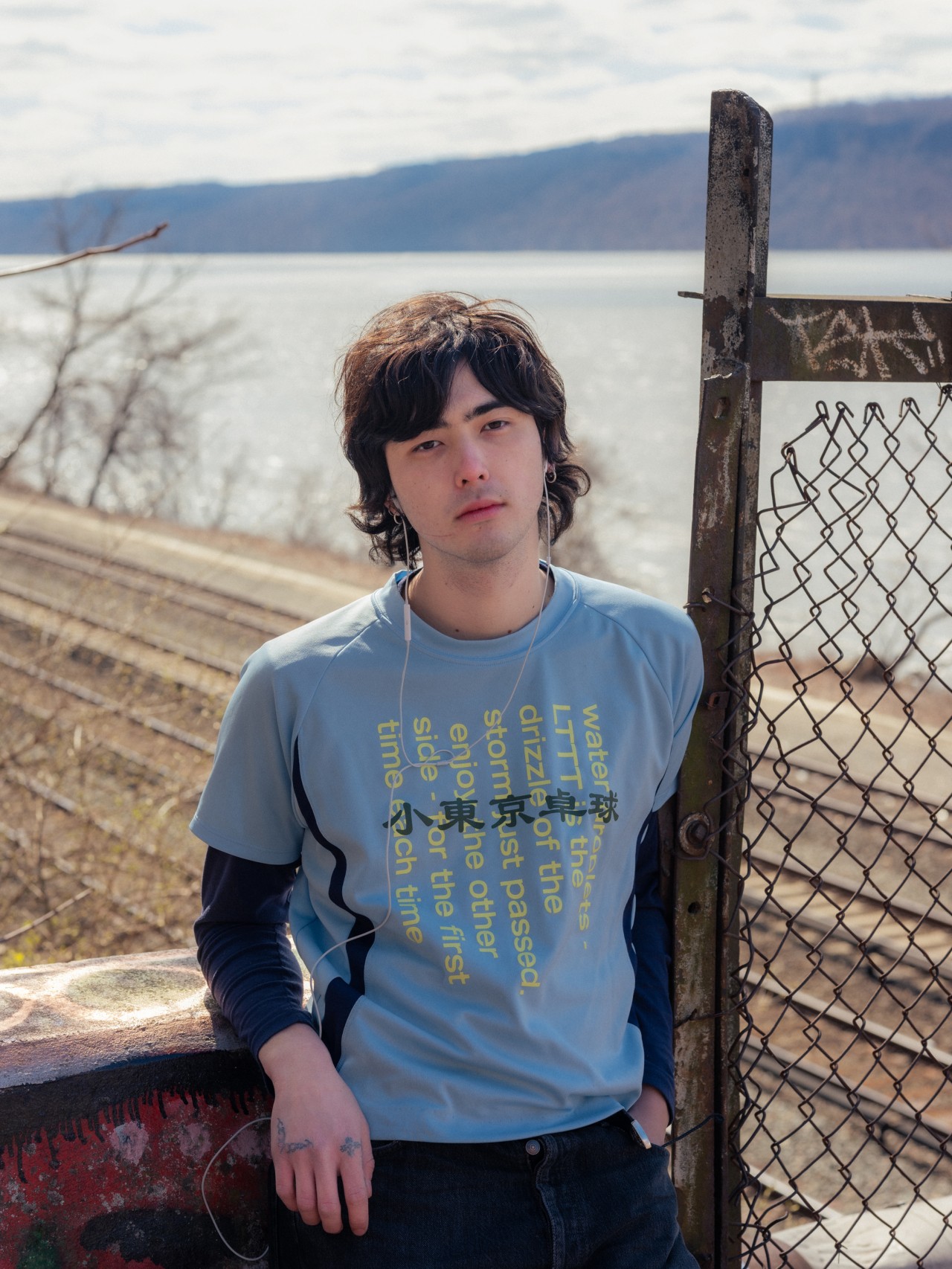 A young Asian American man stands next to a railroad track. The ocean is in the distance.
