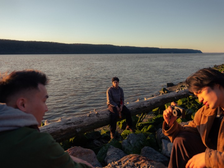 Three young asian men are sitting near the edge of the ocean. One is taking a photo of the other.