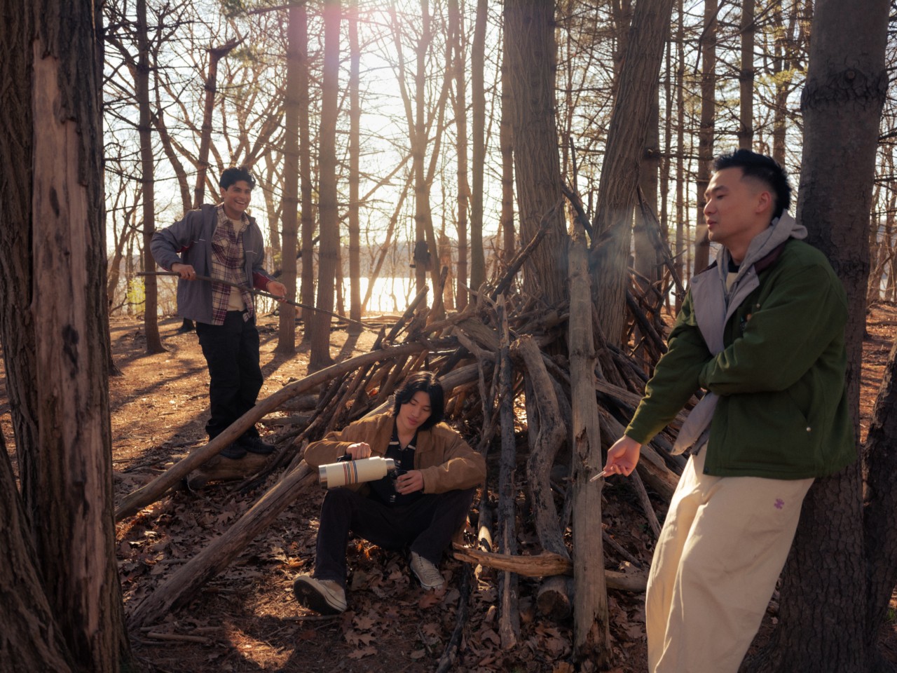 Three young Asian-American men hang out in a forest. One smokes, one dances, and the other sits on the ground while pouring from a canteen.