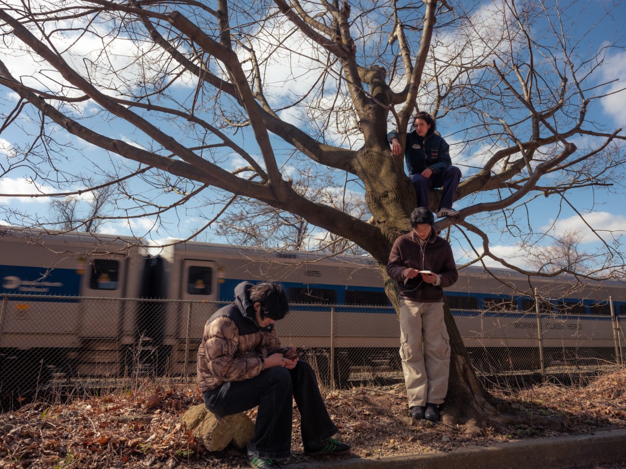 Three young Asian-American men convene next to a railroad track as a train passes by. One sits in a tree, one leans against the tree, and the other sits on a nearby rock.