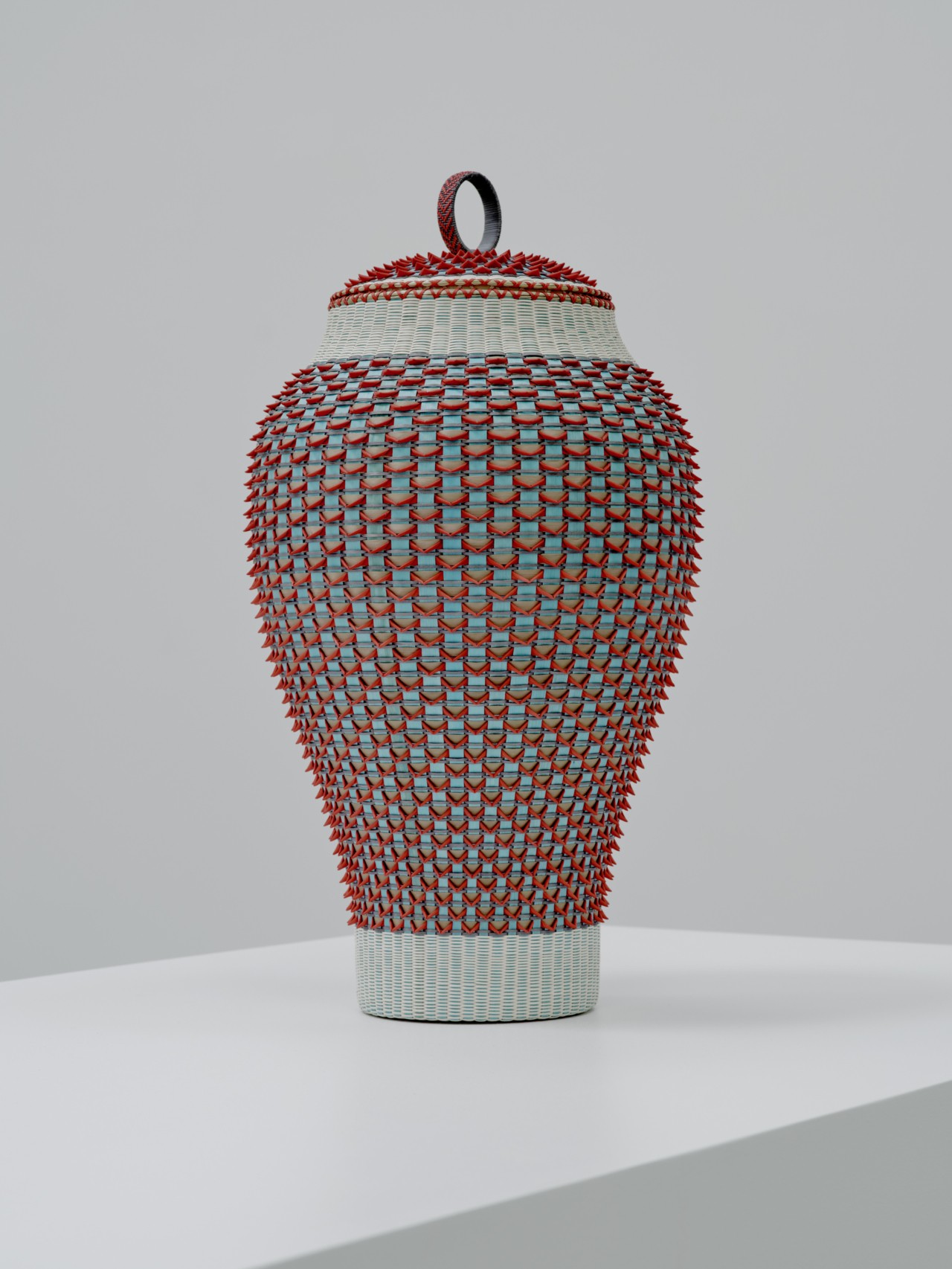 A tall red, black, and blue basket stands on top of a white shelf in a gallery.