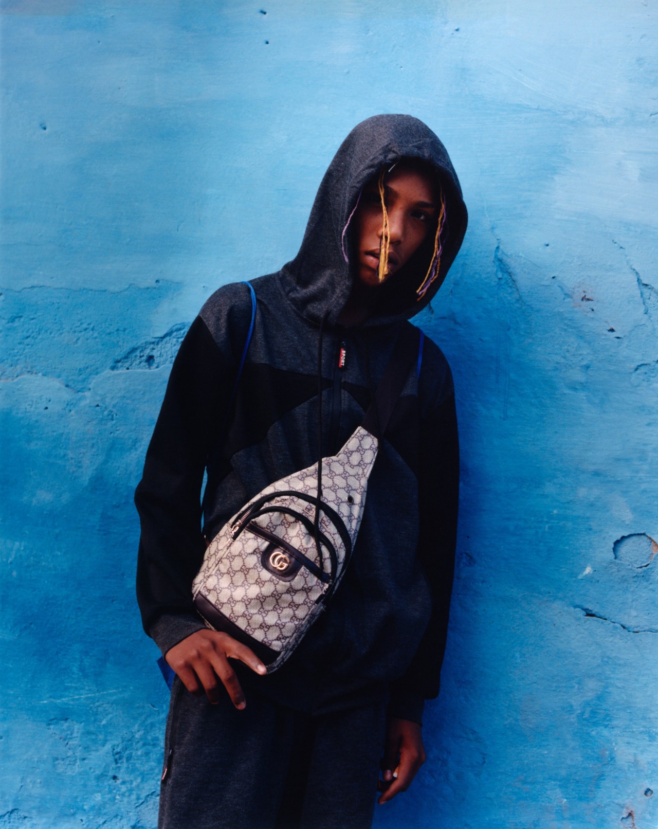 A young Cuban person poses in front of a blue wall. They are wearing a dark blue hoodie and a bag across their chest.