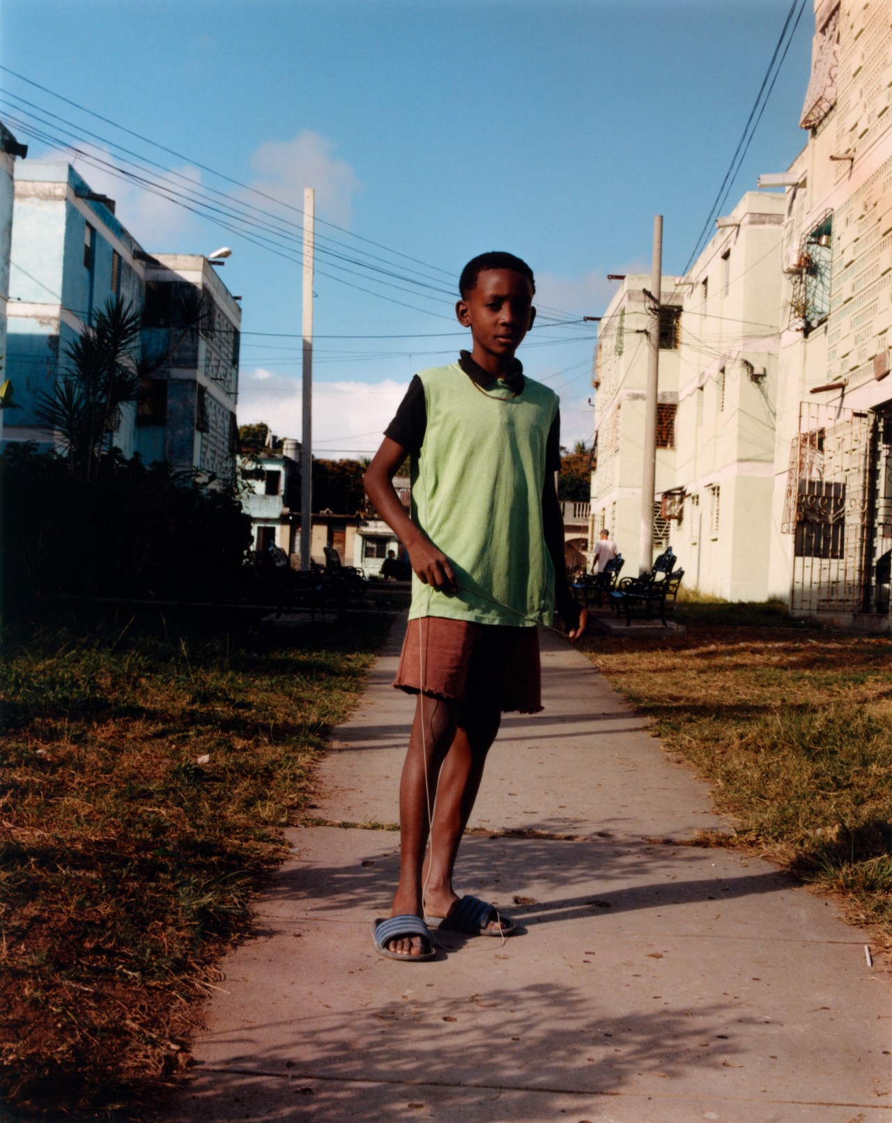 A young boy wearing a bright green shirt, shorts, and sandals stands in the middle of the sidewalk in Cuba.