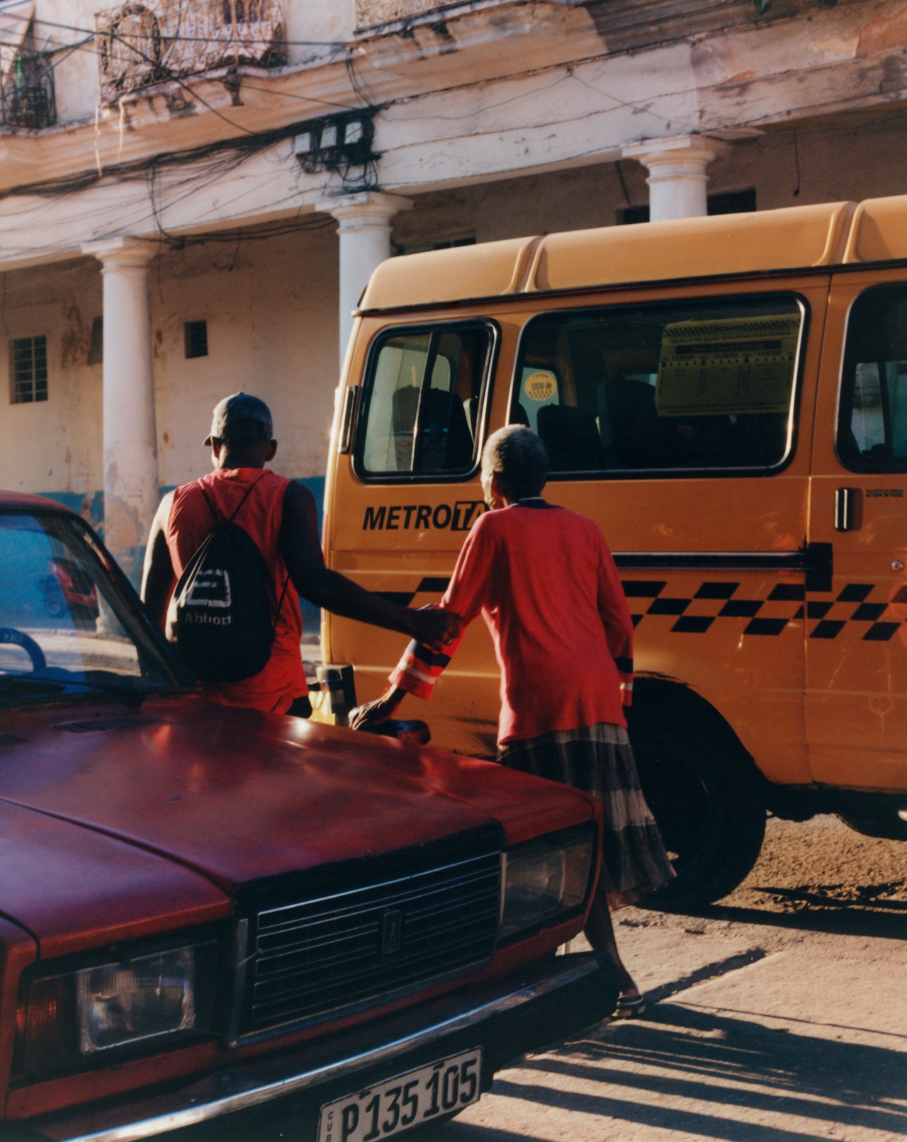 A man helps an older woman cross the street in Cuba as a yellow taxi van rides by.