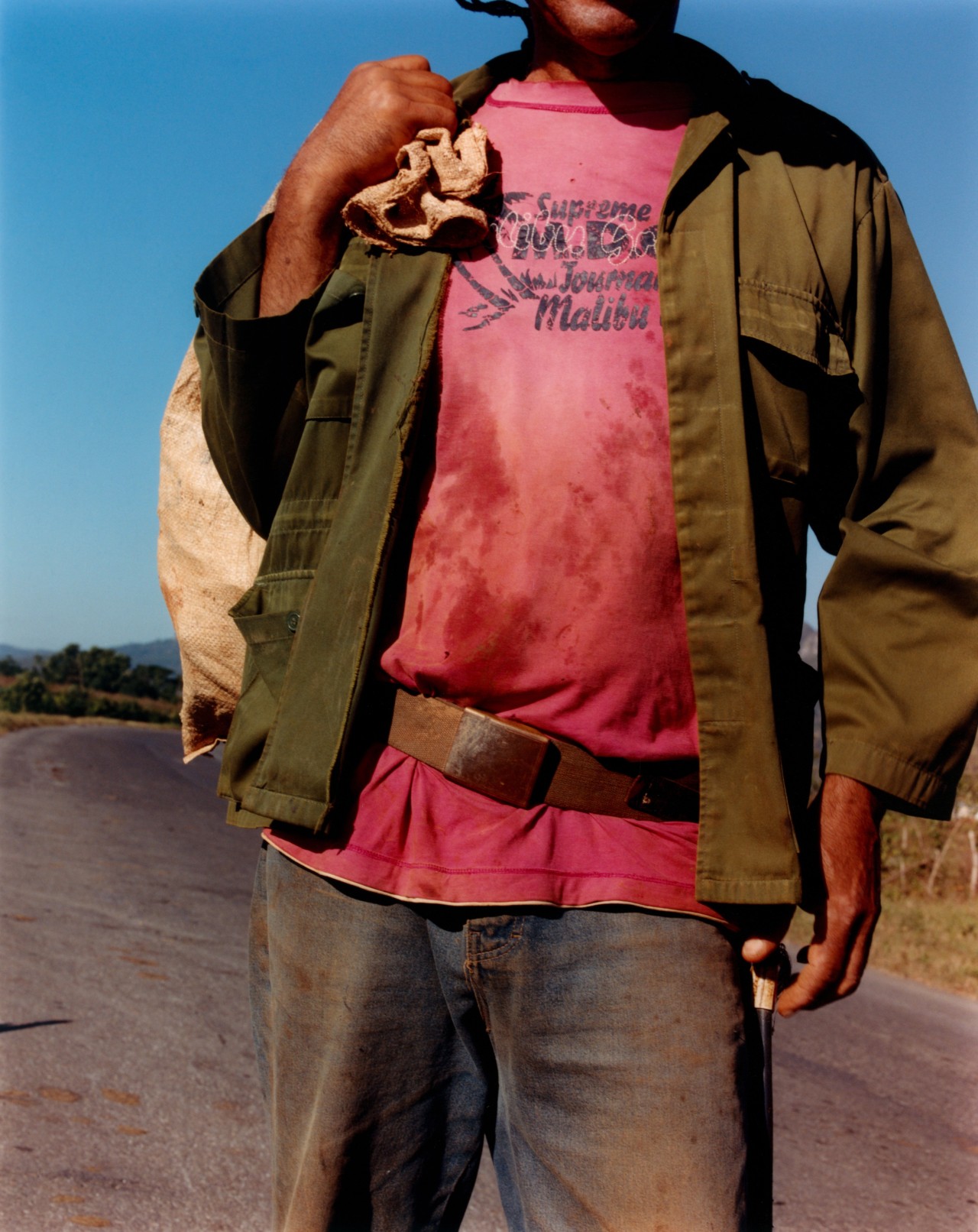 A Cuban person dressed in a red shirt, navy jacket, and jeans poses in the middle of the street while carrying a knap sack over their shoulder.