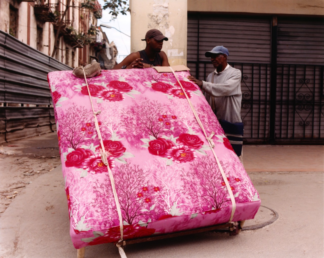 Two Cuban men converse while moving a large pink and red mattress through the streets of Cuba.