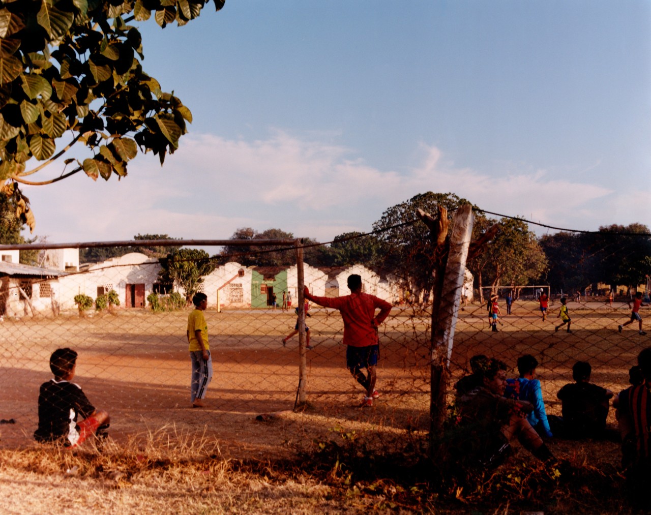Several people watch a soccer game outside from the other side of a fence.