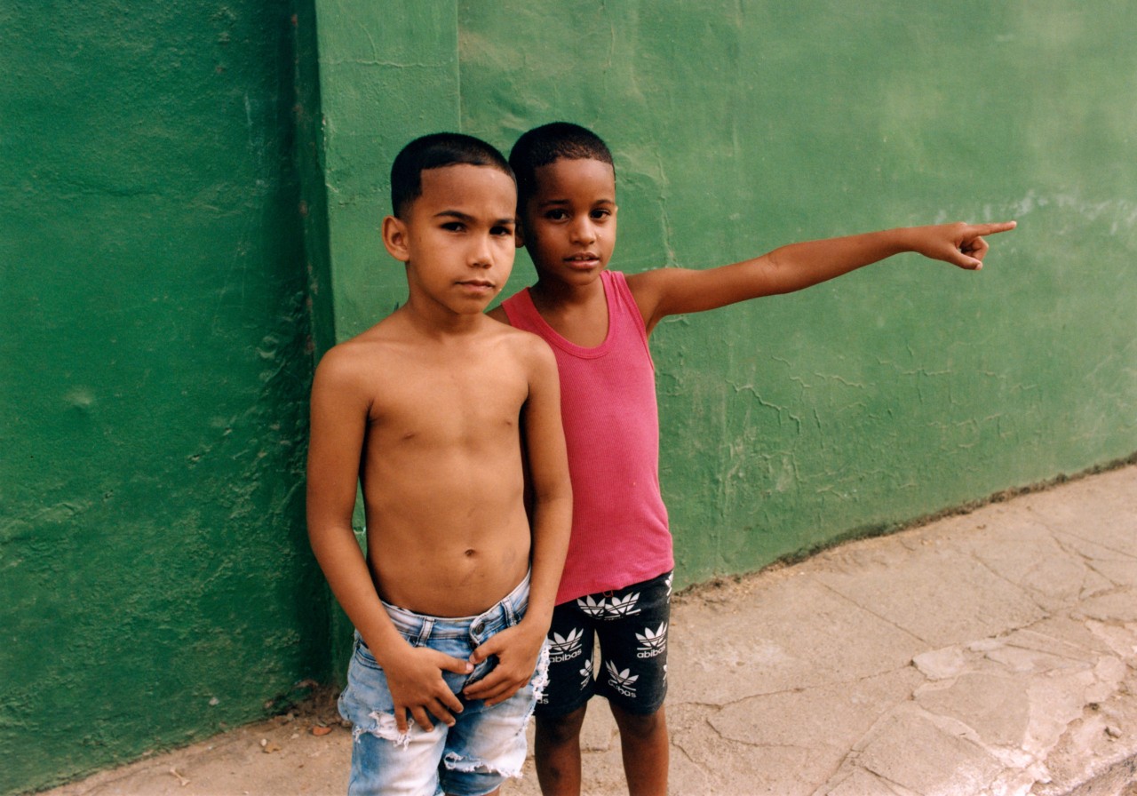 Two young Cuban boys stand in front of a green painted wall. One of them points toward the left.
