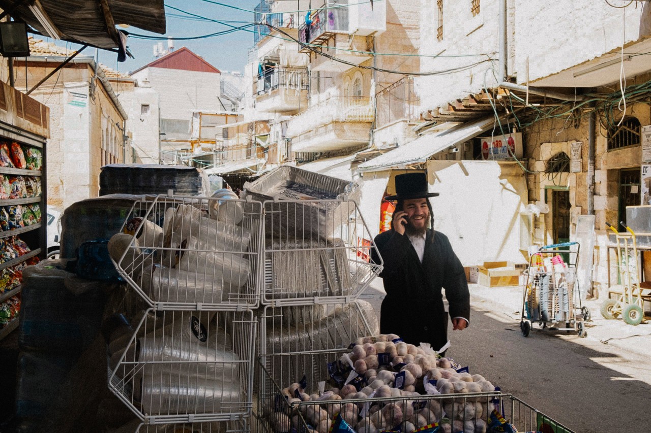 An ultra-Orthodox man chats on the phone while walking through a Jewish neighborhood with plastic vendors around him.