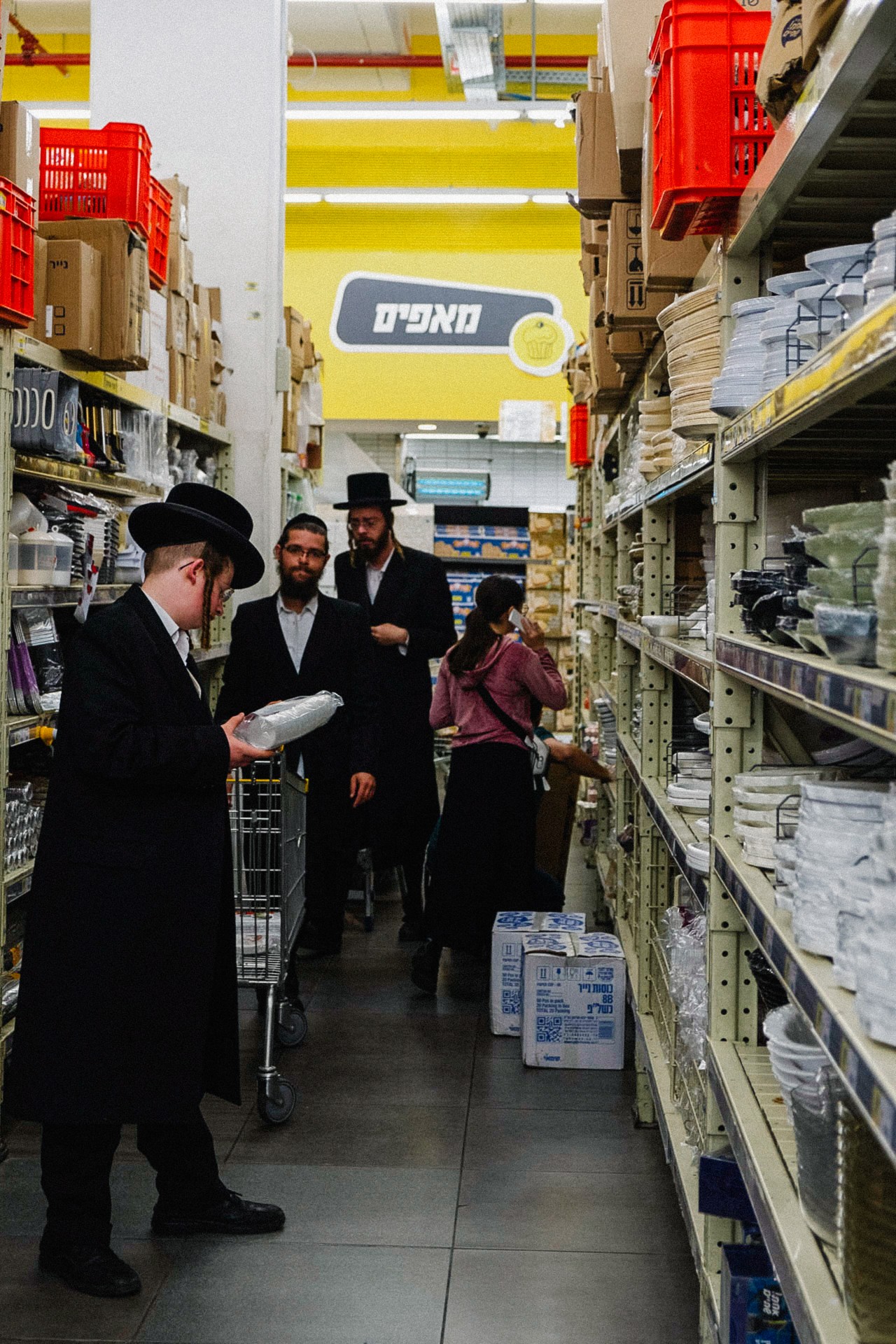 Three ultra-Orthodox Jewish men shop for plastic products in a narrow aisle of the grocery store.