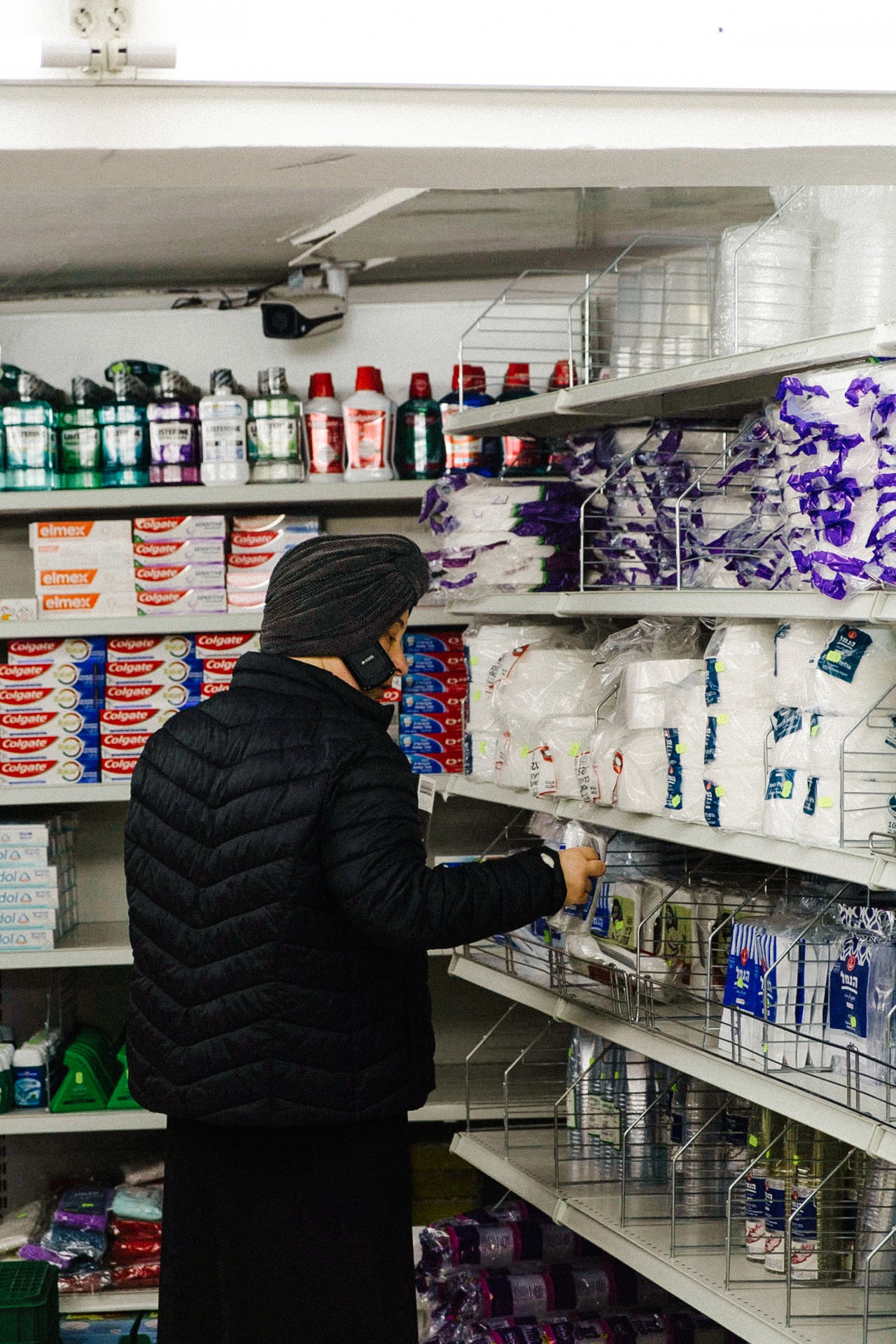 An ultra-Orthodox Jewish man faces away from the camera, toward the plastics section of the grocery store, picking up one of the products and observing it.