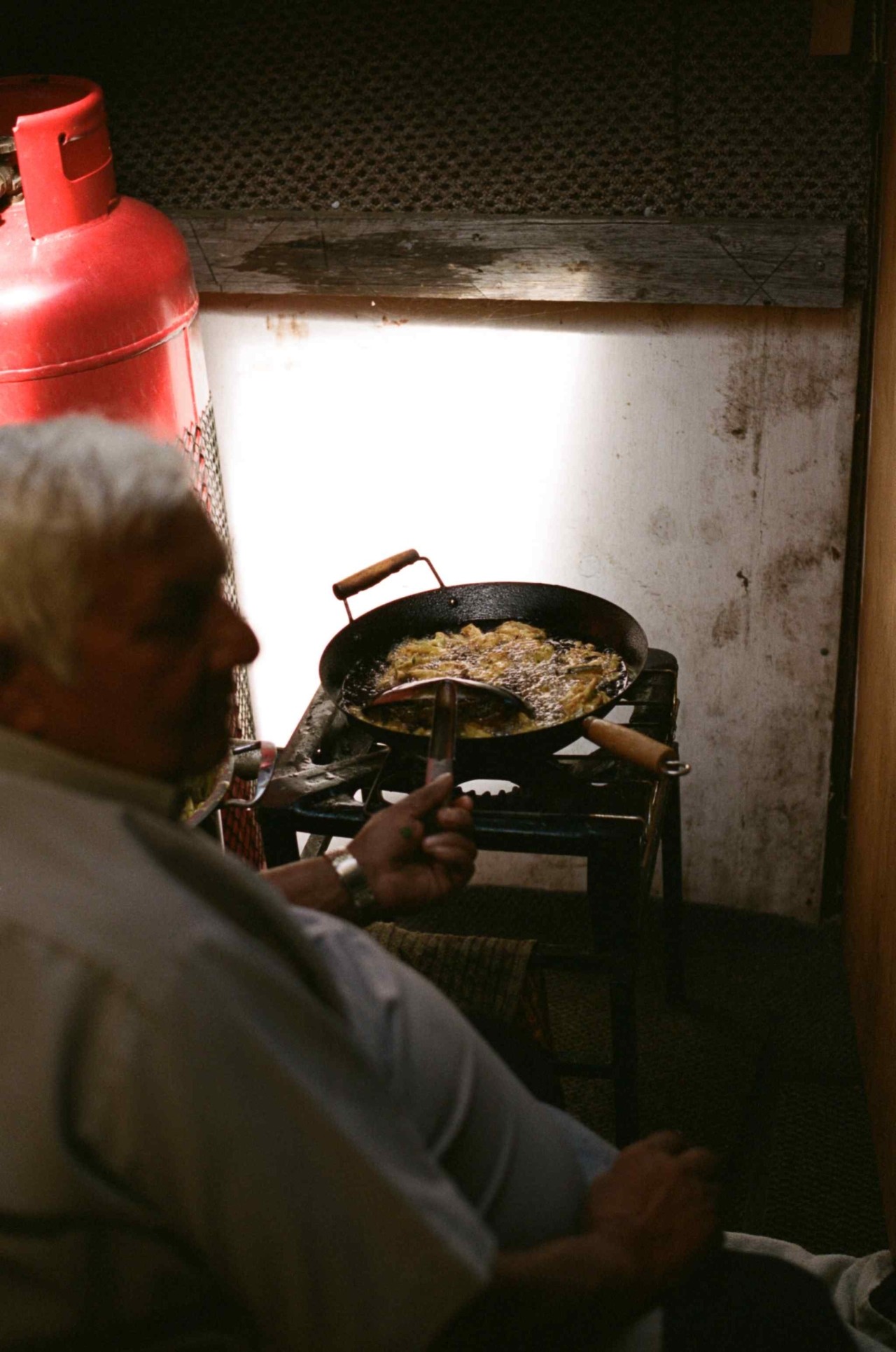 An older man sits down while frying food in small black pan on a small stove.