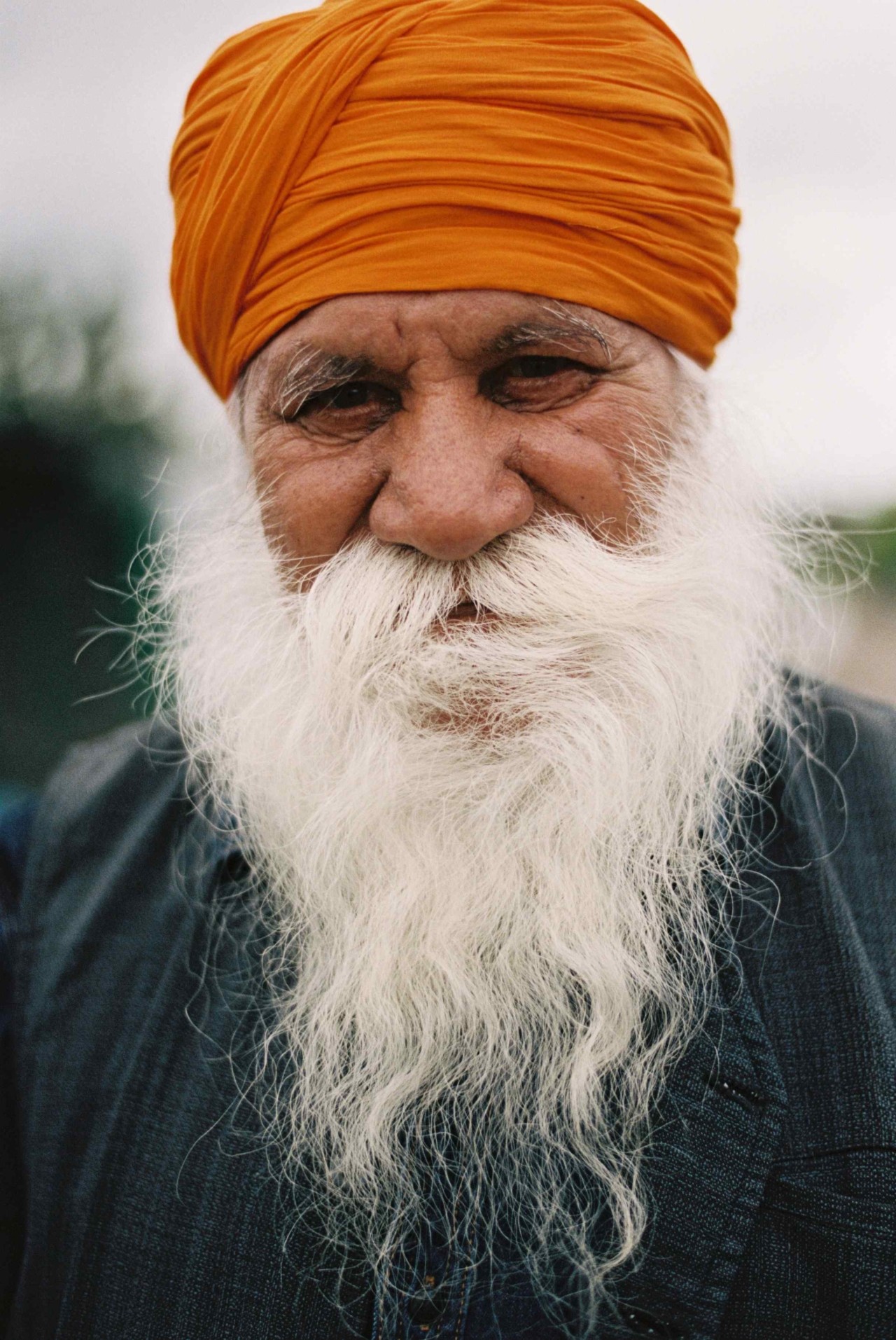 An older Punjabi Sikh farmer with a long white beard and a bright orange head wrap stares into the camera.