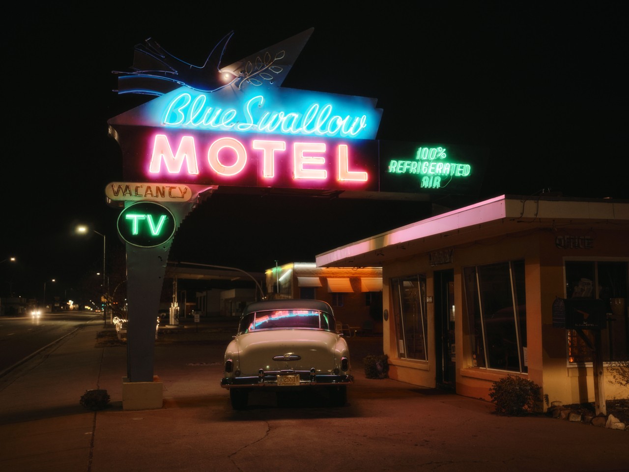 A 1950s looking car is parked right outside a motel with a lit up sign hanging above it. It's night time.