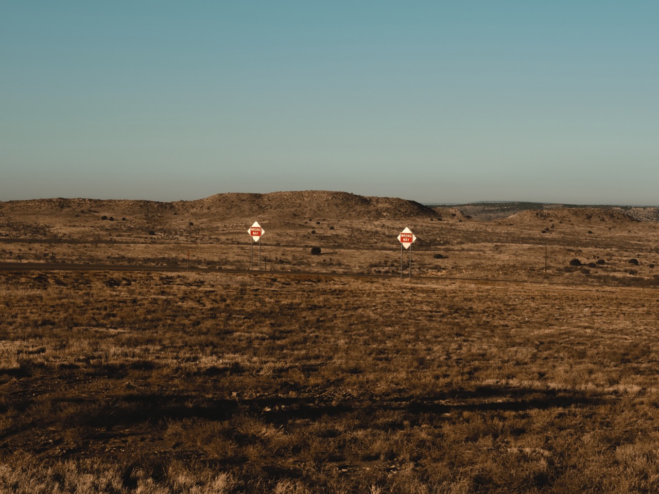 Two lonely signs stand a few feet apart in the distance in a large dirt field.