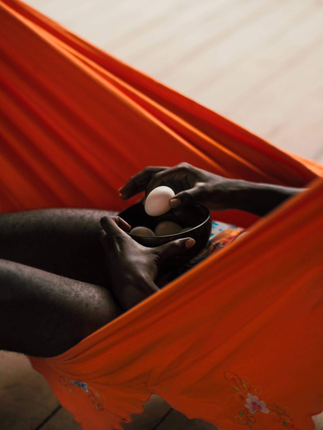 A person lying down in an orange hammock is holding a bowl of eggs. In one hand, they carefully hold a single egg between their fingers.