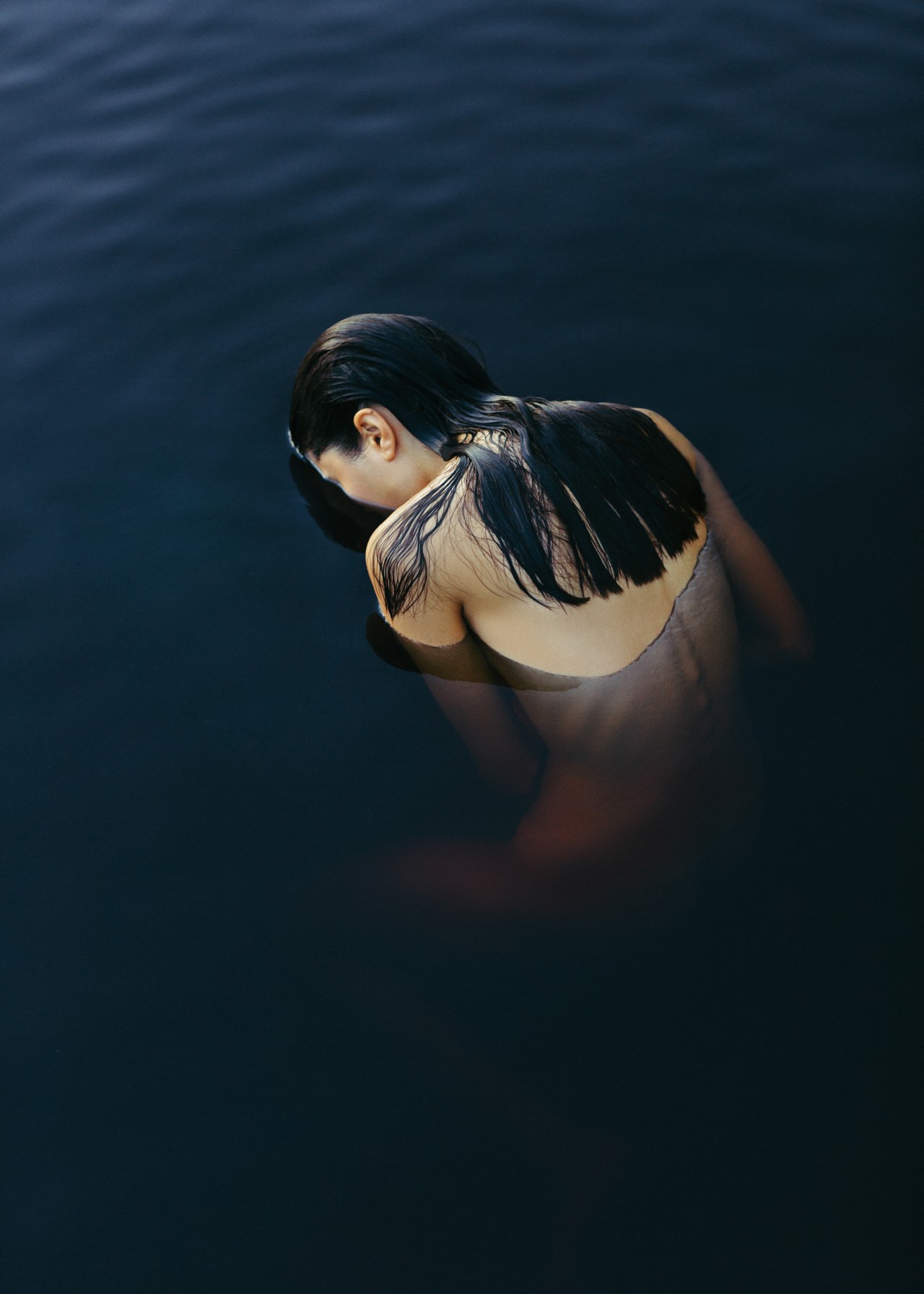 A person with long, jet black hair wearing no clothing faces away from the camera, partially submerging their face in the water in the Amazon rainforest.