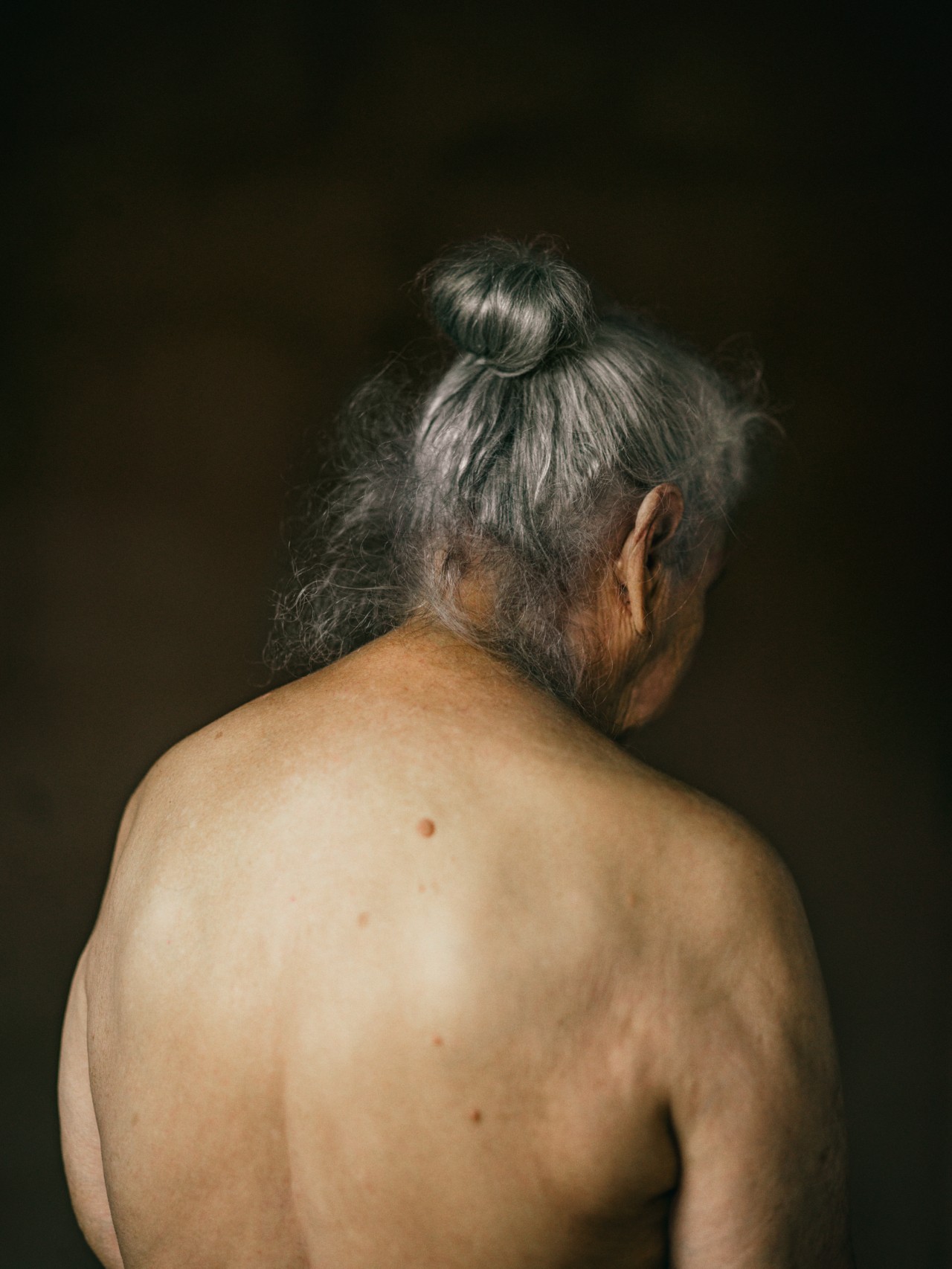 An older person with a gray bun turns their back to the camera. The backdrop is completely black.