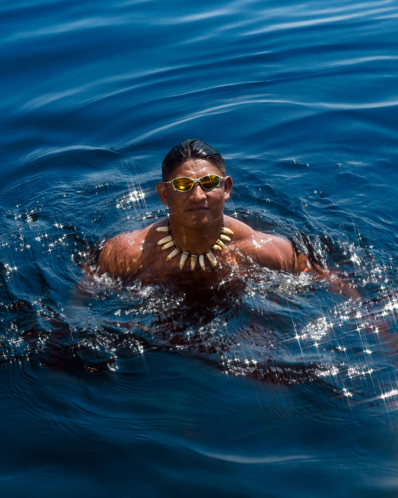 A man wearing sunglasses and a tooth necklace treads in the deep blue waters in the Amazon rainforest.