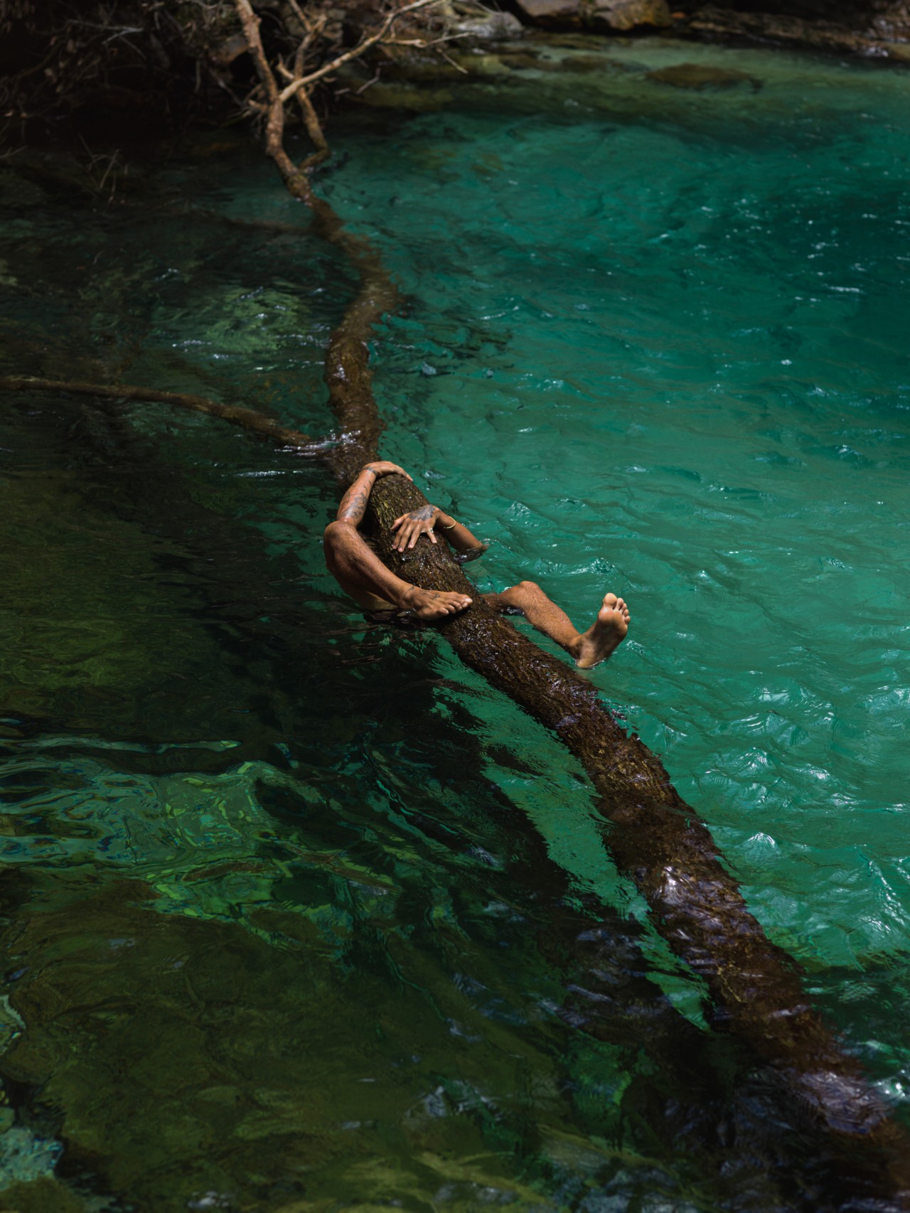 A person's arms and legs are shown straddling a fallen tree in the middle of a blue lagoon in the Amazon rainforest.