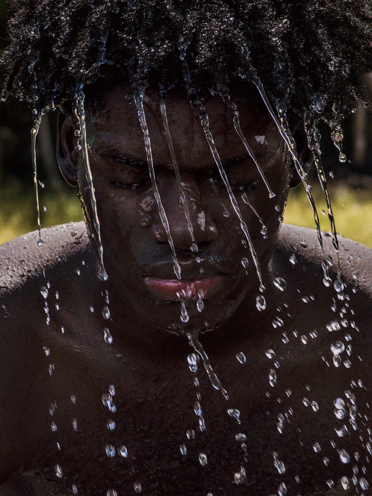 A young man peers down as water streams from his dark, kinky curls.